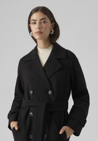Cappotto doppiopetto nero realizzato in misto lana, con ampio colletto, vita con cintura e bottoni grandi. Tessitura liscia, design lungo fino al ginocchio.
