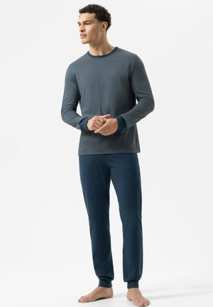 Junger Mann, der barfuß steht und ein blau gestreiftes Langarmshirt sowie eine einfarbige blaue Hose trägt, während er zur Seite auf einem weißen Hintergrund schaut.