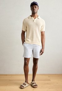 Knit cream polo shirt, light gray textured shorts, and beige sandals. Model wearing a navy cap, standing on a wood floor with a neutral background.