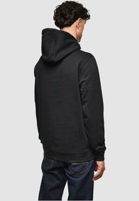 Schwarzer Hoodie mit flacher Strickkapuze, gerippten Bündchen und Saum sowie einer glatten, weichen Textur. Trägt er mit dunklen Jeans.