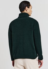 Mann, der von hinten zu sehen ist, trägt einen dunkelgrünen Rollkragenpullover und beige Hosen vor einem schlichten hellen Hintergrund.