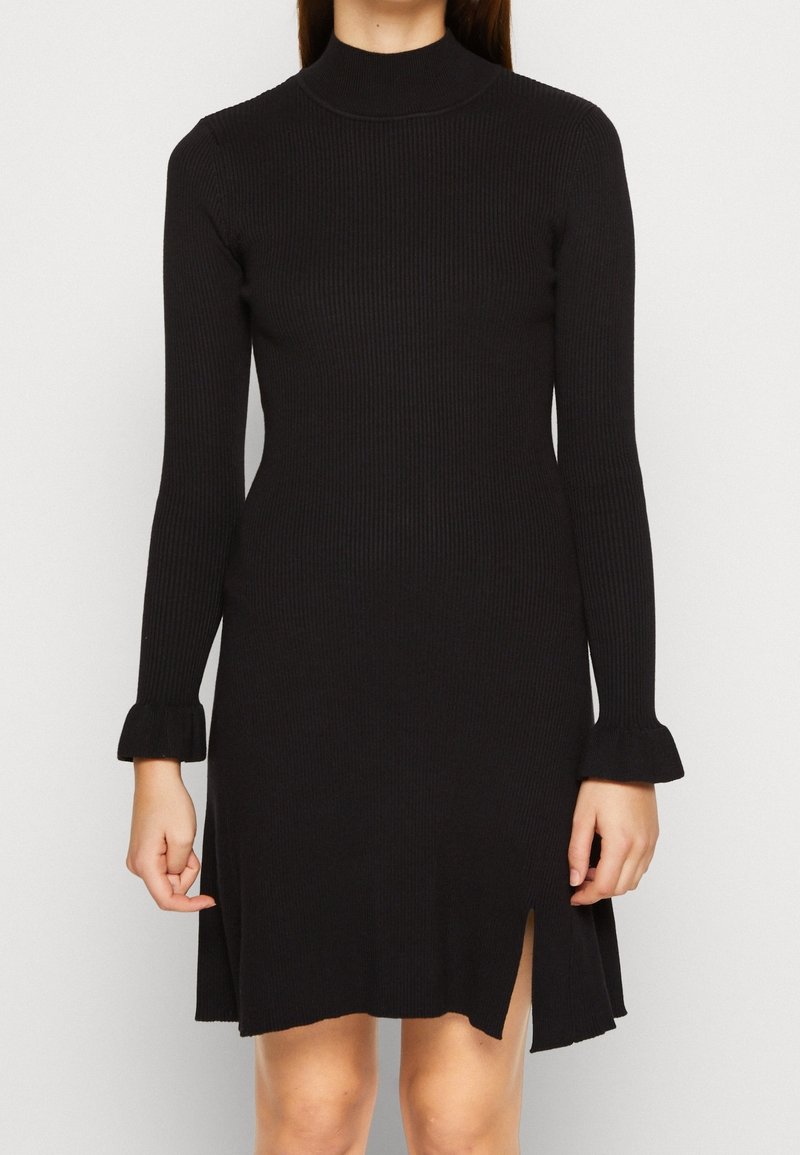 Musta resorineulottu mekko, jossa pitkät hihat, korkea pääntie ja röyhelöiset kalvosimet; sivuhalkiot ja kapea istuvuus.