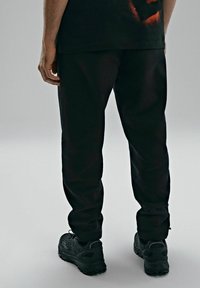 Czarne joggerki o zwężanym kroju z gładką teksturą, elastycznym pasem i zamkami błyskawicznymi przy kostkach, w połączeniu z czarnymi butami sportowymi.