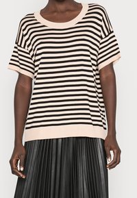 Persoon in een beige en zwart horizontaal gestreept kortmouwshirt, gecombineerd met een zwarte geplooide rok, staande tegen een effen achtergrond.