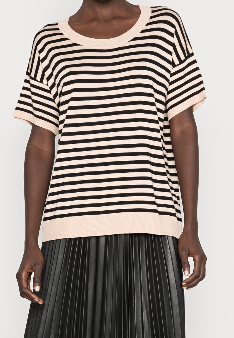 Persoon in een beige en zwart horizontaal gestreept kortmouwshirt, gecombineerd met een zwarte geplooide rok, staande tegen een effen achtergrond.