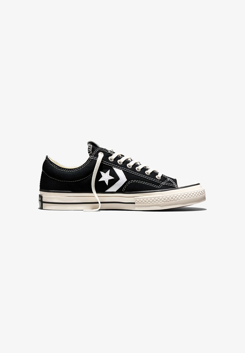 Zapatilla negra de lona de corte bajo con logo blanco de estrella y flecha, cordones blancos y suela de goma color crema con puntera texturizada.