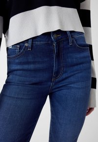 Jean en denim bleu foncé à coupe ajustée, avec poches avant, fermeture à boutons visible et coutures discrètes sur les coutures.
