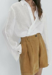 Weiße Leinenbluse mit langen Ärmeln und Kragen, kombiniert mit hochgeschnittenen, hellbraunen Leinen-Shorts mit einer Knopfdetails vorne.