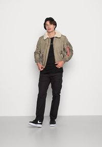 Chaqueta bomber caqui con un cuello de piel sintética, puños acanalados y bolsillos con cremallera, combinada con pantalones cargo negros y zapatillas negras.
