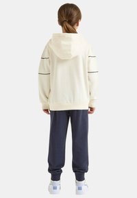 Felpa con cappuccio crema con strisce nere, abbinata a pantaloni della tuta blu navy. Scarpe da ginnastica bianche con dettagli blu. Tessuto liscio e vestibilità rilassata.