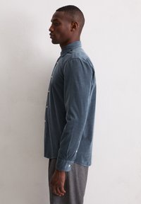 Junger Mann, der seitlich steht und ein langärmliges blaues Hemd und graue Hosen trägt, vor einer schlichten weißen Wand.