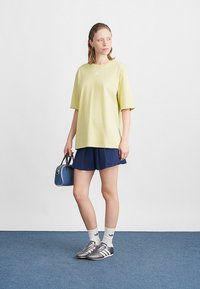 Ανοιχτό κίτρινο Oversized T-shirt, ναυτίες πλισέ σορτς, γκρι αθλητικά παπούτσια και λευκές κάλτσες με σχέδιο. Μια μικρή μπλε τσάντα προσθέτει λεπτομέρεια.