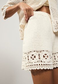 Bílé háčkované šortky mají texturovaný vzor se zvlněným lemem a kruhovými výřezy. Pas je elastický pro maximální pohodlí.