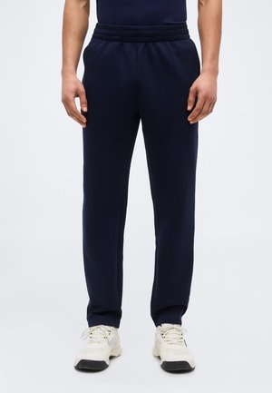 Άνδρας που φοράει ναυτικό μπλε παντελόνι φόρμας και λευκά αθλητικά παπούτσια, στέκεται μπροστά σε απλό ανοιχτόχρωμο φόντο.