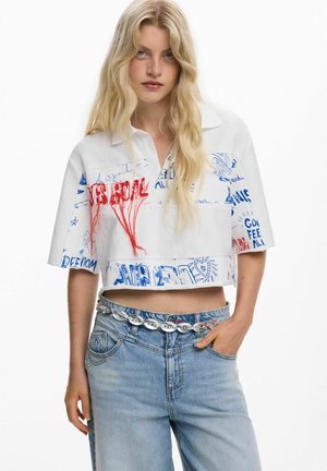 Femeie blondă purtând o cămașă albă scurtă cu text de graffiti roșu și albastru, asortată cu blugi de culoare albastru deschis și o curea argintie cu zale.