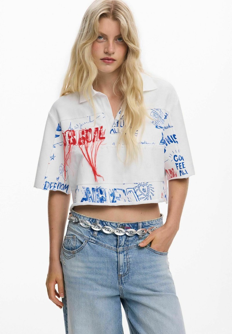 Mujer rubia con una camiseta blanca corta con texto de graffiti en rojo y azul, combinada con jeans azul claro y un cinturón de cadena plateada.
