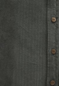 Strukturiertes olivgrünes Cordstoffgewebe mit vertikalen Rippungen, kombiniert mit braunen Knöpfen an einer Seite. Dezente Markenkennzeichnung auf den Knöpfen.