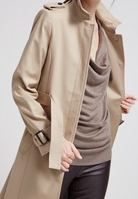 Beige trenchcoat met hoge kraag, schouderepauletten en riem. Gelaagd bruin topje eronder, gecombineerd met donkere, strakke broek.