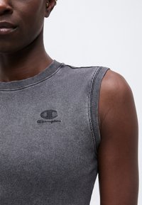 Κοντινό πλάνο ενός ατόμου που φοράει αμάνικη γκρι ριγέ μπλούζα με μικρό μαύρο λογότυπο Champion στο στήθος.