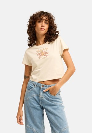 Junge Frau mit lockigem Haar trägt ein leichtes, kurzes T-Shirt mit Pferdeprint und lockere hellblaue Jeans, posiert mit der Hand in der Tasche.