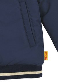 Die marineblaue Jacke verfügt über eine glatte Textur, eine Seitentasche und gerippte Bündchen mit cremefarbenen Streifen. Sie enthält ein gelbes Etikett mit einem Bärenlogo.