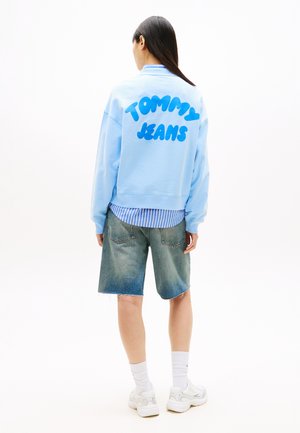 Γυναίκα με μακριά σκουρόχρωμα μαλλιά που φοράει ανοιχτό μπλε φούτερ με τη λέξη "Tommy Jeans" στην πλάτη, τζιν σορτς, λευκές κάλτσες και λευκά αθλητικά παπούτσια.