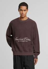 Brązowy sweter z dzianiny z ocieplanymi rękawami i dołem. Na przodzie znajduje się biały haftowany logo z napisem "Another Cotton Established 2023".
