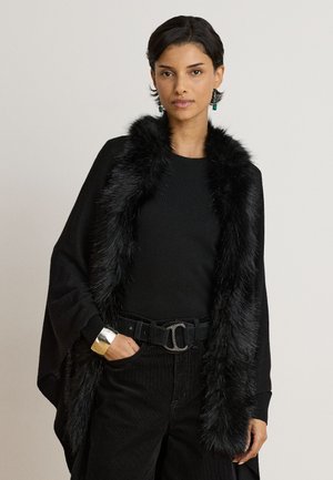 Donna con top nero e pantaloni, scialle nero con bordatura in pelliccia, bracciale in argento e orecchini verdi su sfondo neutro.