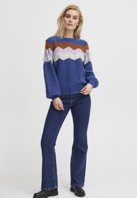  Blauwe gebreide trui met zigzagpatroon in bruin, wit en paars, gecombineerd met een flared donkere jeans en zwarte laarzen.