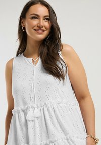 Vit ärmlös klänning med ögla tyg, dragbandsstängning vid halsen och en flerlagers volangslåt med tofsar.