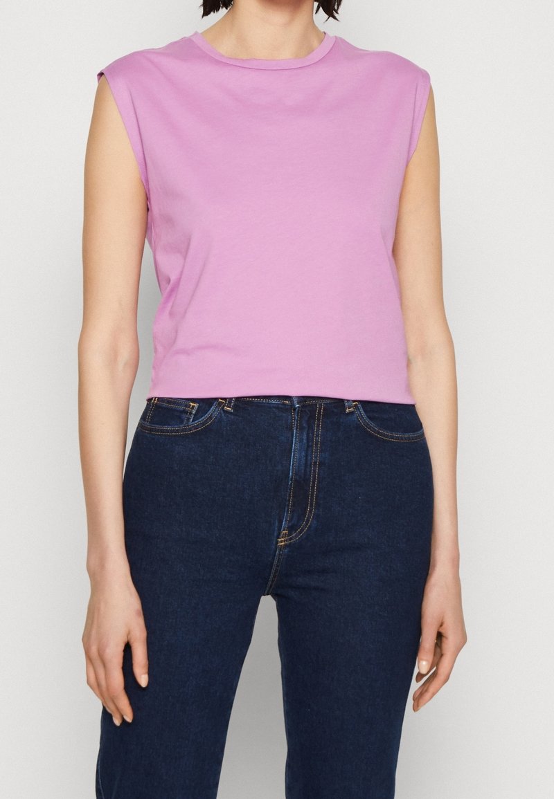 Bezruký krátký top z světle růžové bavlny, má kulatý výstřih a jemné akcenty na ramenou, je kombinován s tmavě modrými džíny.