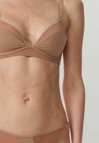 Femme portant un soutien-gorge couleur chair et une culotte assortie, montrant son torse et son bras contre un fond clair.