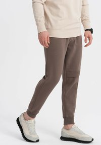 Brune joggerbukser med en struktureret stof, tætsiddende pasform, sidelomme til cargo og elastisk talje, suppleret med lyse sneakers.