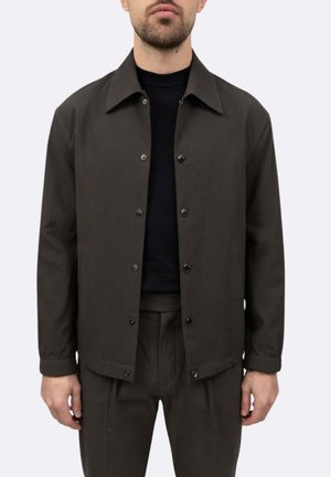 Uomo che indossa una giacca verde oliva scuro con bottoni sopra una camicia nera e pantaloni verde oliva scuro abbinati su sfondo chiaro uniforme.