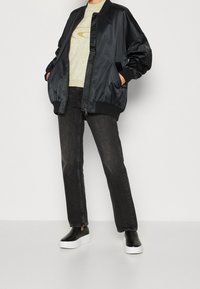 Veste bomber noire avec poignets côtelés, fermeture éclair devant et poches latérales. Associée à un pull vert clair et un jean en denim foncé. Chaussures noires à enfiler.