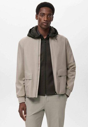 Beige lightweight jacket with a dark green hood, front zip closure, and two patch pockets. Smooth texture and relaxed fit.