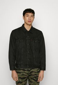 Jachetă din denim negru cu guler, buzunare frontale și închidere cu nasturi. Purtată peste pantaloni cu model de camuflaj.