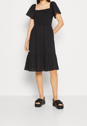 Femme portant une robe noire mi-longue avec des manches bouffantes et un décolleté carré smocké, associée à des sandales compensées noires.