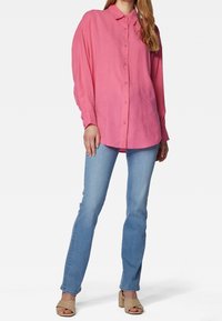 Camicia rosa a maniche lunghe con colletto, dalla vestibilità rilassata e texture morbida, abbinata a jeans blu chiaro e sandali con zeppa beige.