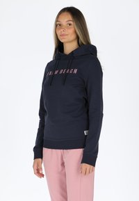 Felpa blu marina con testo rosa "PALM BEACH". Presenta cappuccio con lacci, polsini e orlo a coste. Indossata con pantaloni rosa chiaro.
