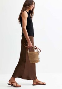 Top sans manches noir, jupe maxi marron, sandales à lanières marron, portant un sac à main en osier avec une sangle blanche, présentant une finition lisse et des lignes épurées.