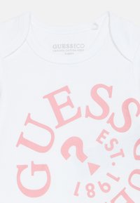 Baltos organinės medvilnės bodysuit'as su dideliu rožiniu grafiniu tekstu, kuriame užrašyta "GUESS" ir metų skaičius "1981" įvairiais šriftais ir dydžiais.