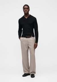 Chemise noire à manches longues côtelée avec un col doux, associée à un pantalon plissé beige clair et des chaussures noires, créant un look formel.