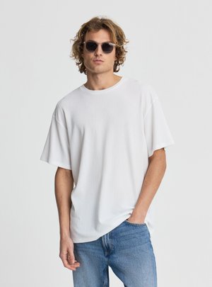 Giovane uomo con capelli ricci che indossa occhiali da sole neri, una maglietta bianca larga e testurizzata e jeans blu, posando con una mano in tasca.