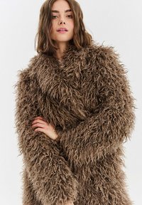 Hnědý huňatý kabát s chlupatou texturou, nadměrným střihem a širokými rukávy. Materiál působí plyšově a teple.
