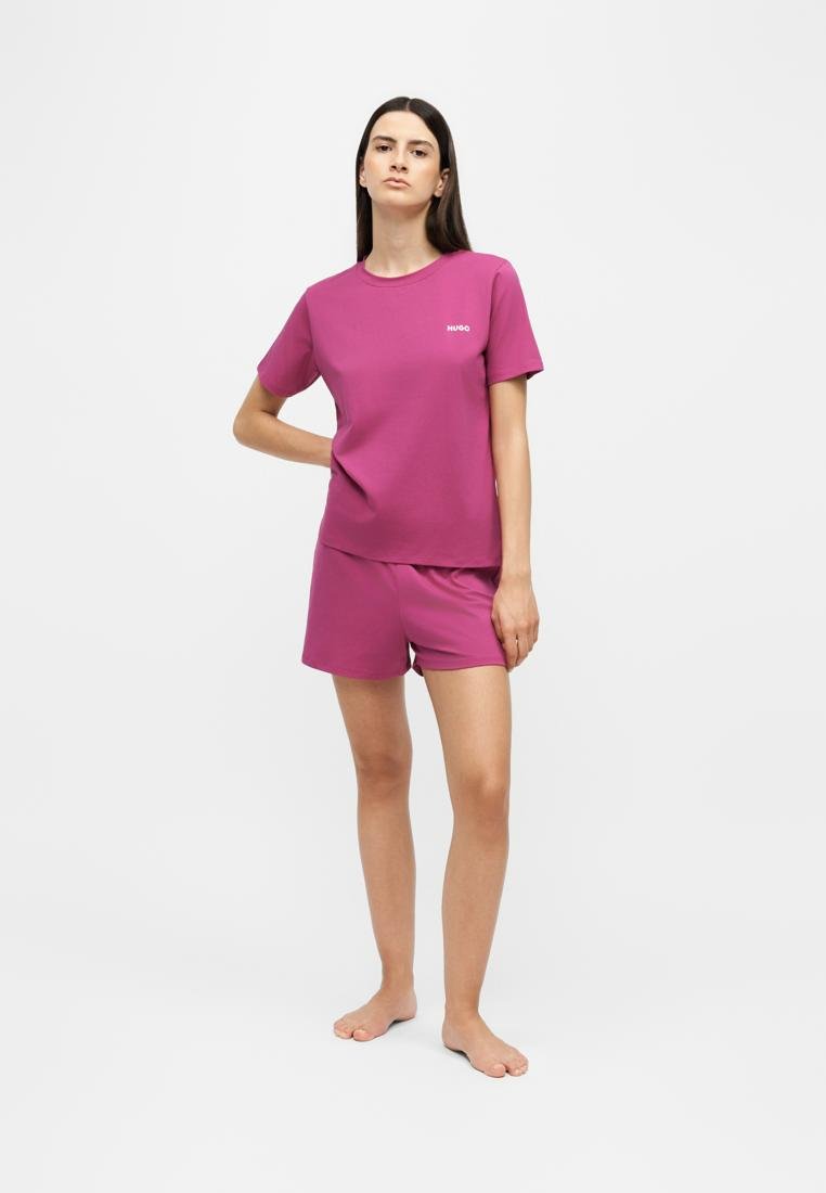 Femme debout avec une main sur la hanche, portant un ensemble violet à manches courtes et short, sur un fond blanc uni.