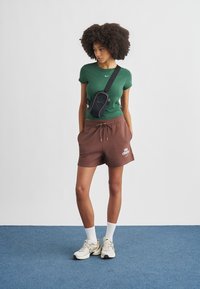 Πράσινο εφαρμοστό T-shirt, καφέ σορτς με κορδόνι, μαύρη τσάντα χιαστί, λευκό λογότυπο Nike, λευκές κάλτσες γυμναστηρίου και λευκά αθλητικά παπούτσια με γκρι λεπτομέρειες.