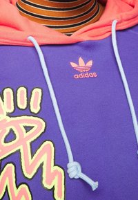 Fialová mikina s růžovým akcentem na kapuci, která má velký grafický design a modré tkaničky. Obsahuje růžové logo adidas.