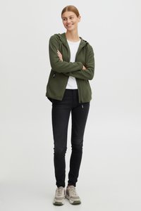 Felpa verde oliva con zip e cappuccio regolabile, indossata sopra una T-shirt bianca, abbinata a jeans neri skinny e sneakers beige.