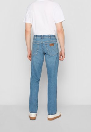 Homme portant un jean bleu clair droit, un t-shirt blanc à manches courtes et des chaussures blanches avec des semelles marron, debout devant un fond uni.